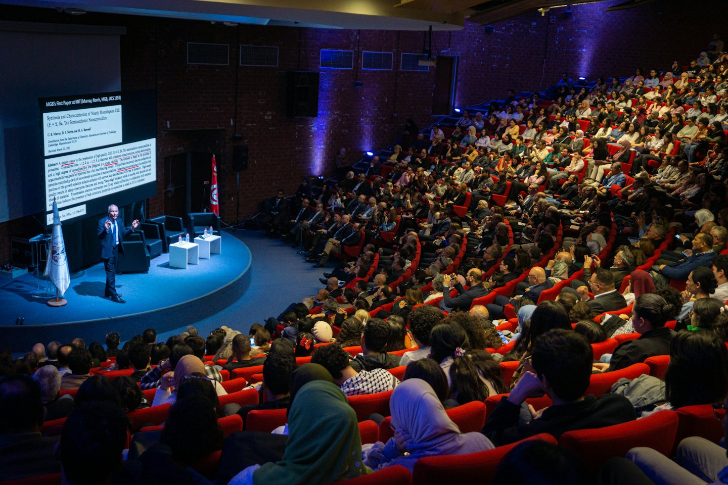 Man presents a scientific paper on a blue stage to an audience.